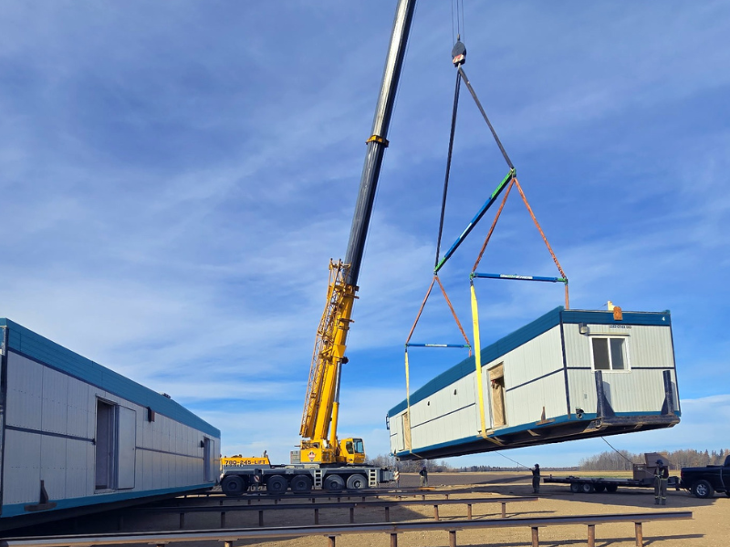 Pre-fab modular camp hoisted by a crane with a blue sky and dirt foreground